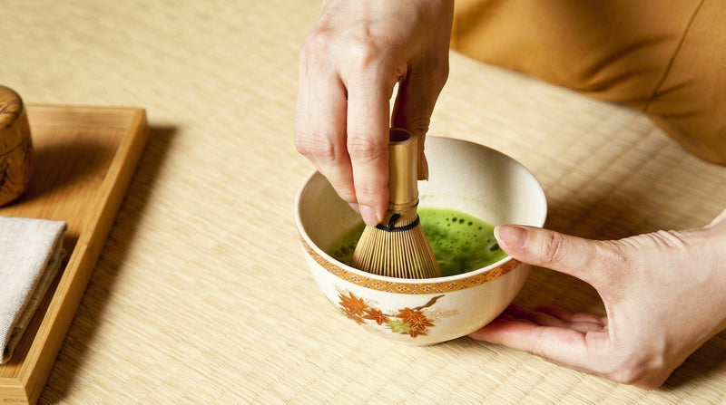Des mains fouettent le matcha avec un chasen dans un bol blanc orné de feuilles d'érable, posé sur un tatami